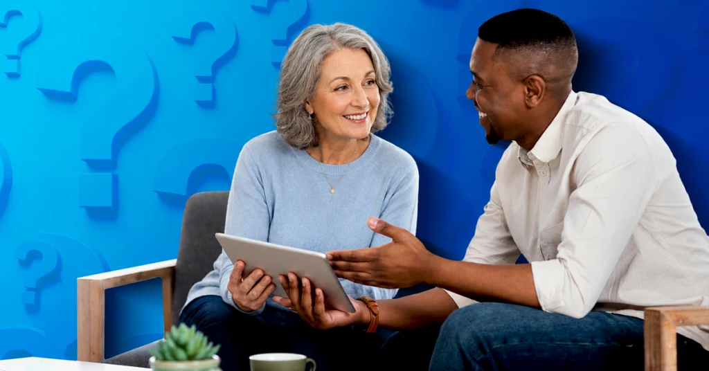 An image of a smiling older woman with a tablet having something explained to her by a kind young black man. The background is blue with question marks.