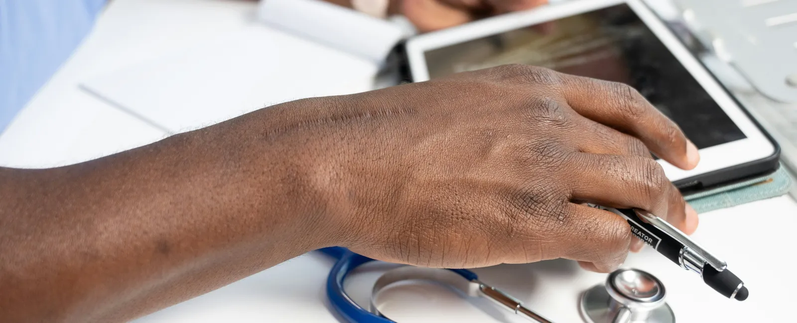 Close-up of a doctor using a HIPAA compliance software enabled tablet with a stethoscope and pen on a white desk, showing medical consultation.